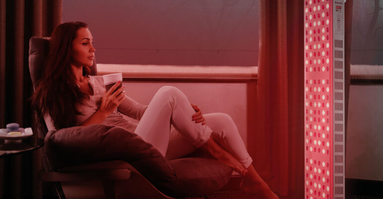 Beautiful young woman at home sitting on modern chair in front of window, relaxing in her living room and drinking coffee or tea